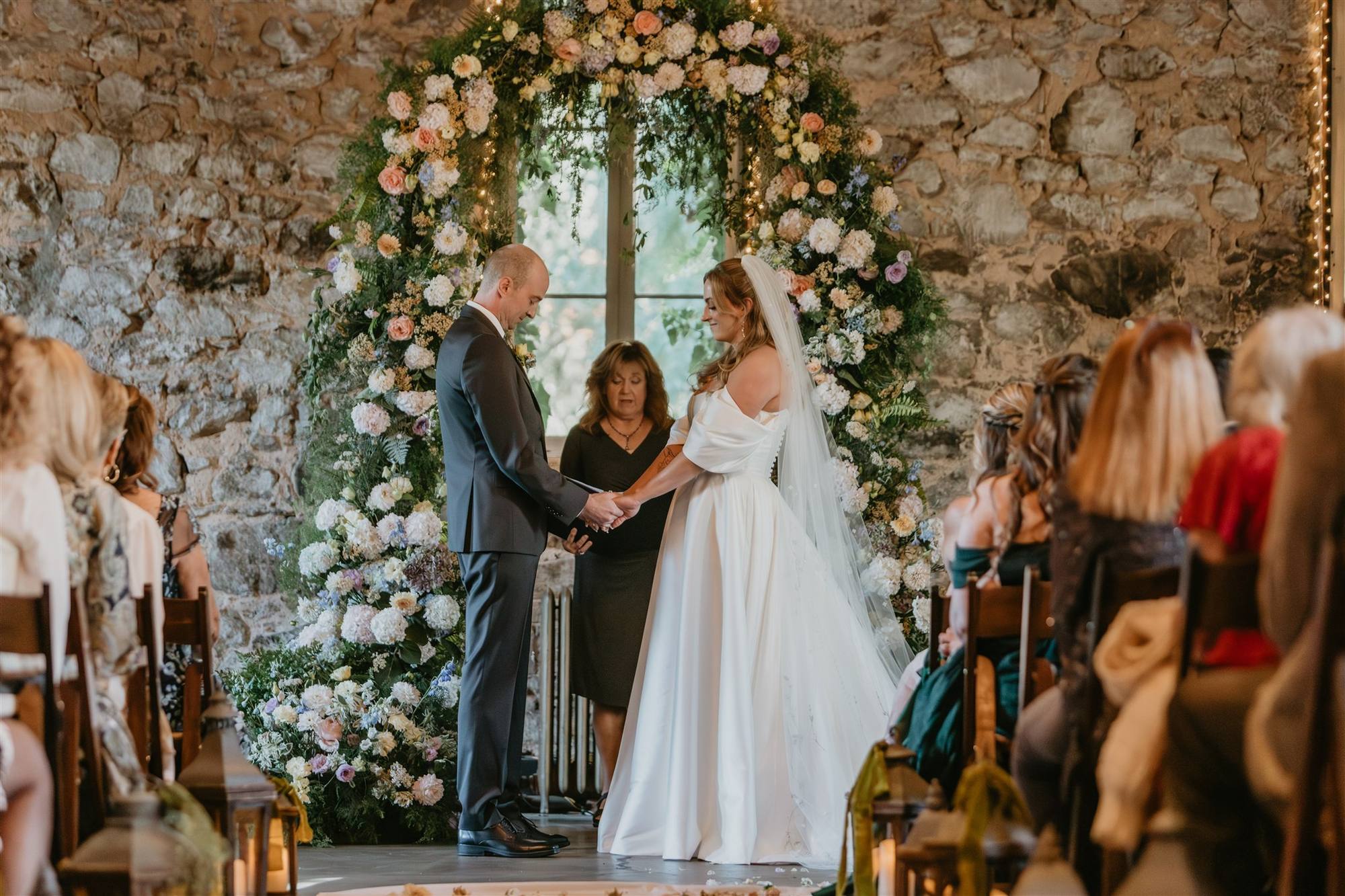 Stone-walled ceremony room: a couple holding hands before their officiant under a towering arch of blush, peach and cream blooms framing a tall window
