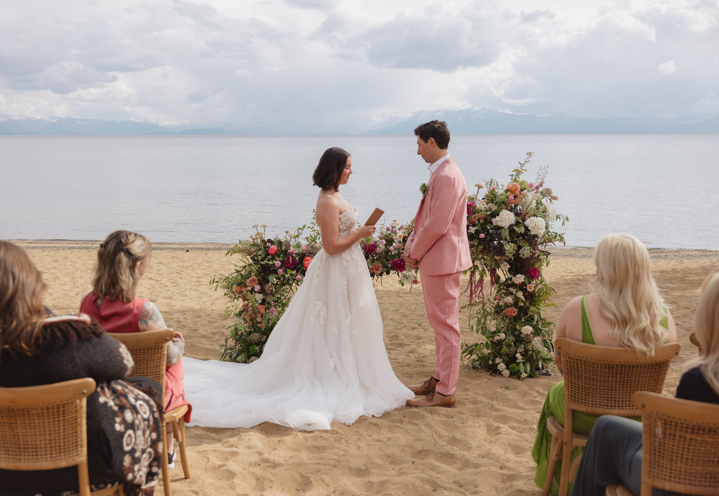 Lake Tahoe beach ceremony: bride reading her vows to a groom in a pink suit between two coral and burgundy floral installations, guests seated on the sand
